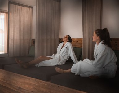 Zwei Frauen entspannen in weißen Bademänteln auf einer Liege in einem Spa-Raum