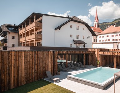 Hotel mit Außenpool und Liegestühlen vor Bergkulisse und Kirche