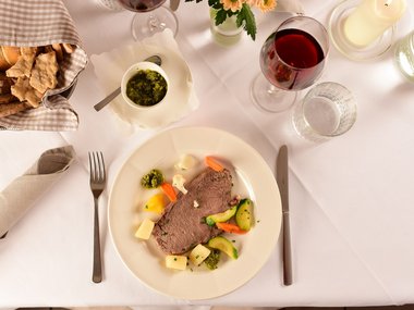 Plate with beef, vegetables, pesto, bread basket and red wine on white tablecloth