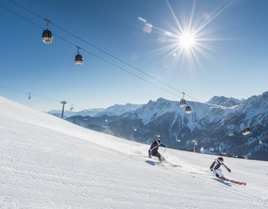 Zwei Skifahrer fahren auf schneebedeckter Piste unter Sesselliften bei sonnigem Himmel