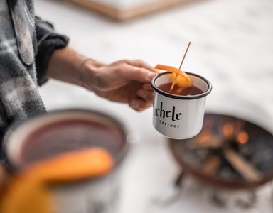 Zwei Hände halten Tassen mit heißem Getränk und Orangenschale am Lagerfeuer