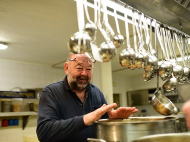 Uomo anziano riscalda le mani sopra una grande pentola in cucina con mestoli appesi