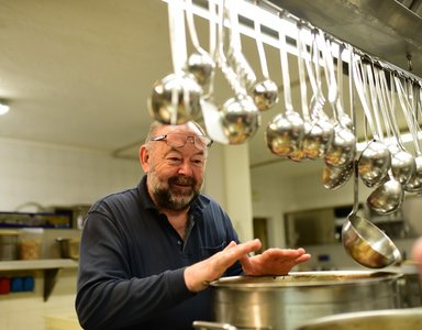 Älterer Mann wärmt Hände über großem Topf in Küche mit gehängten Schöpfkellen