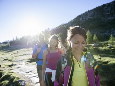 Drei Wanderer auf einem sonnigen Bergweg mit Rucksäcken und Sportkleidung