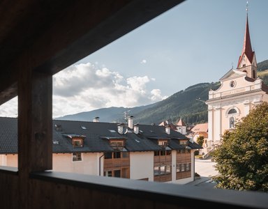 Blick aus Holzrahmen auf Häuser und Kirche vor bewaldeten Bergen